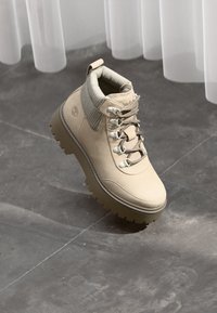 Beige lærretsekkstøvler med tykk såle, polstret krage og metalløse, plassert mot en grå steingulv med sheer gardiner.