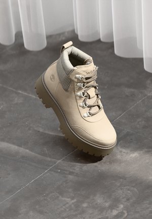 Beige leather hiking boot with thick lug sole, padded collar, and metal eyelets, set against a gray stone floor and sheer curtains.