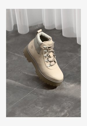 Bota de senderismo de cuero beige con suela gruesa, cuello acolchado y ojales metálicos, situada sobre un suelo de piedra gris y cortinas transparentes.