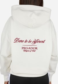 Sudadera blanca de gran tamaño con capucha. En la parte trasera presenta texto bordado en rojo: "Atrévete a ser diferente", "PEGADOR", "Susurros de Frío". Textura suave.