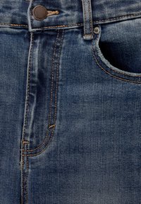 Gros plan de la ceinture d’un jean en denim bleu avec un bouton métallique, une boucle de ceinture et une poche avant avec coutures visibles.