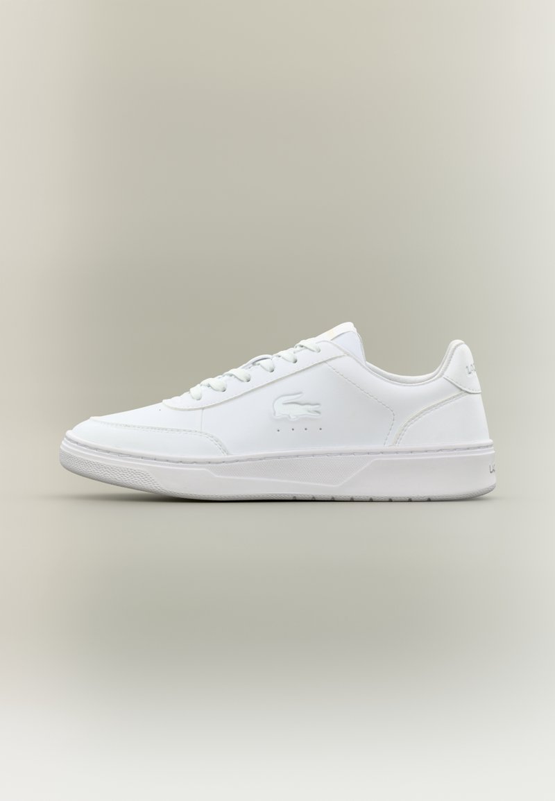 Basket basse blanche à lacets à l'avant, logo crocodile embossé sur le côté et semelle texturée sur fond neutre.