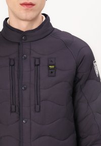 Veste matelassée marine avec un motif à ondulations, fermetures à pression, détails avec cordon de serrage et une patch de la marque sur la poitrine gauche.