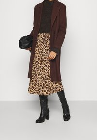 Brauner langer Mantel über einem strukturierten dunklen Pullover, kombiniert mit einem Leopardenmuster-Rock und schwarzen knielangen Stiefeln. Hält eine schwarze Handtasche.