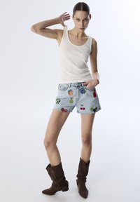 Shorts di denim con ricami di frutta colorata, di colore azzurro chiaro. Abbinati a un top fitted bianco e stivali marroni larghi.
