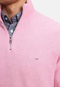 Pull zippé rose clair avec un motif de tricot texturé, comprenant un petit logo brodé et une chemise bleue à motifs en dessous.