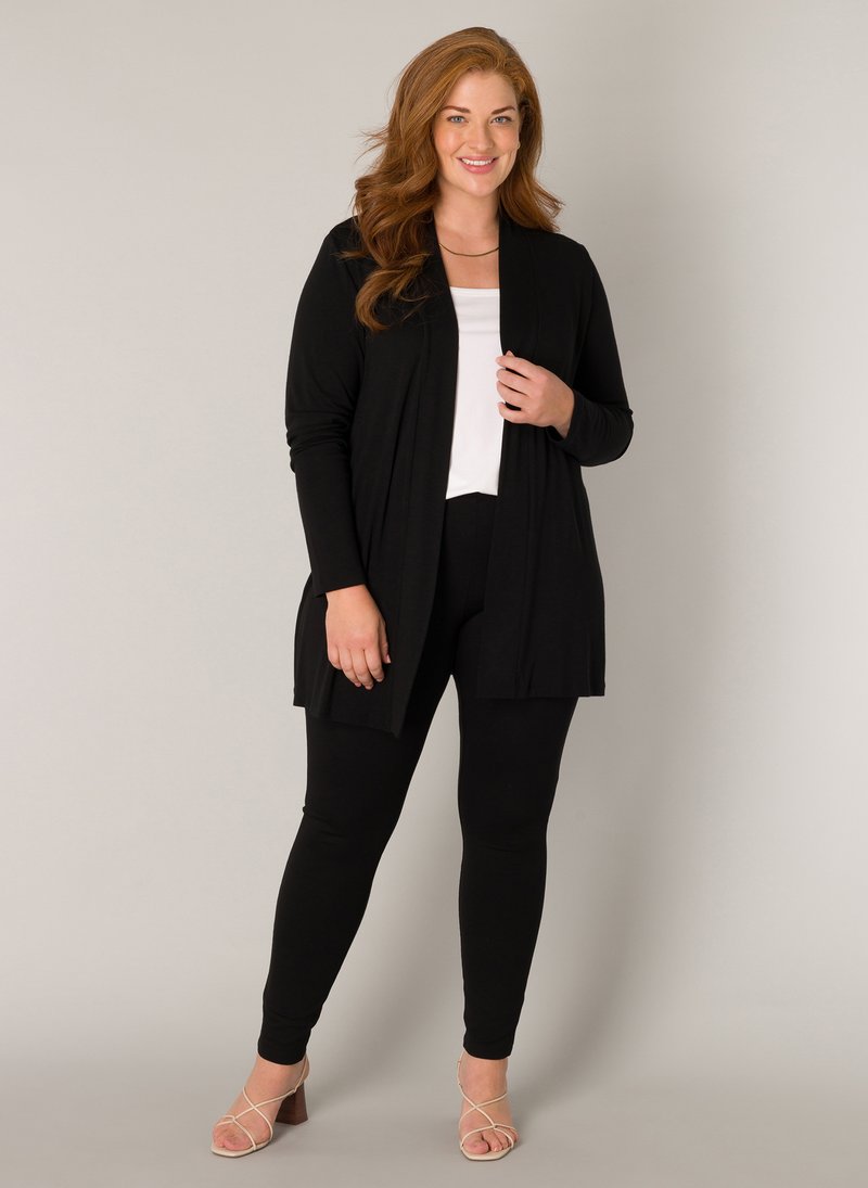 Zwarte cardigan met open voorkant en lange mouwen, gecombineerd met strak zwarte leggings en beige sandalen met bandjes. Witte top eronder.