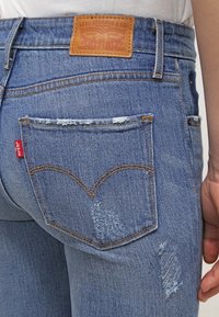 Vaqueros de mezclilla en un azul claro desgastado con un parche de cuero de Levi's, bolsillos traseros prominentes y una etiqueta roja; texturizados con detalles de decoloración y deshilachado.