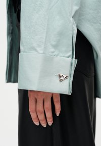 Mano con uñas largas junto a un puño de camisa azul claro abrochado con un gemelo triangular plateado, pantalones negros de fondo.