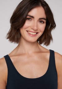Top sin mangas de color azul marino con escote redondeado. Tela de algodón suave con tirantes delgados. El modelo tiene el cabello castaño hasta los hombros y una sonrisa sutil.