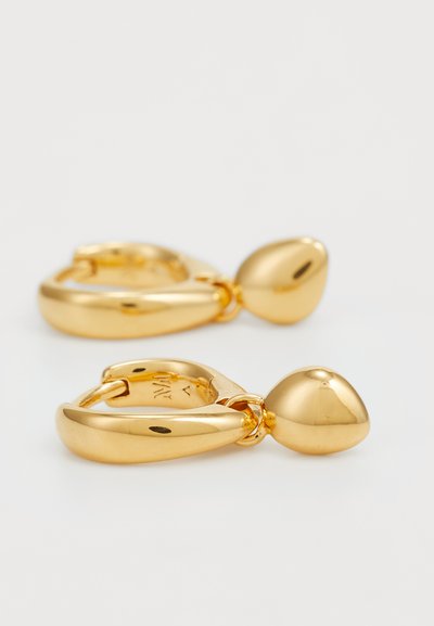 Boucles d'oreilles abstraites en ton or avec une finition polie, présentant un design en forme de goutte d'eau et des bords lisses. Comprend un maillon de connexion circulaire.