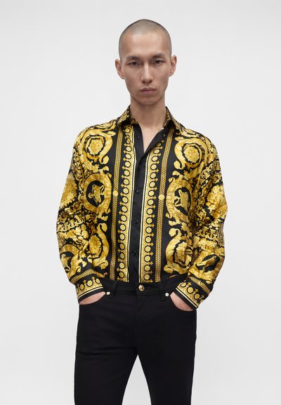 Homme portant une chemise à manches longues noire et dorée ornée, et un pantalon noir, les mains dans les poches, sur fond blanc.