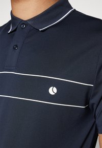 Námornícky polo tričko s bielymi pruhmi, zapínacím golierom a okrúhlym logom na ľavej hrudi. Vyrobené z texturovanej tkaniny. Krátke rukávy.