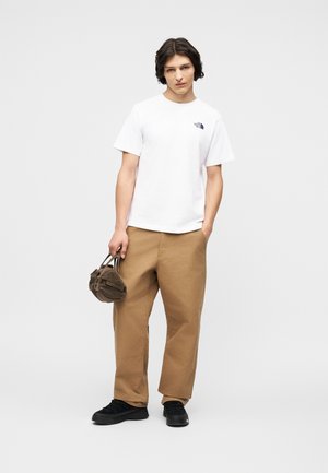 Junge Person steht und trägt ein weißes T-Shirt mit Logo, weite hellbraune Hose, schwarze Schuhe, hält eine kleine braune Tasche in einer Hand, andere Hand in der Tasche.