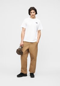 Νεαρός φοράει λευκό μπλουζάκι με λογότυπο, χαλαρά καφέ παντελόνια, μαύρα παπούτσια, κρατώντας μια μικρή καφέ τσάντα στο ένα χέρι, ενώ το άλλο χέρι είναι στην τσέπη.