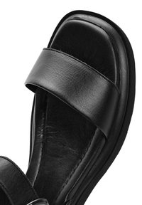 Sandalias de cuero negro con una correa ancha a través del pie, suela plana y una textura lisa y mate. Punta redondeada y diseño minimalista.