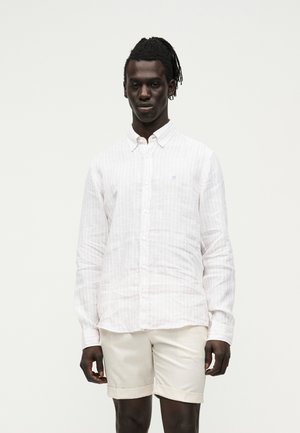 Mann mit Dreadlocks, der ein weißes langärmeliges Hemd und beige Shorts trägt und vor einem schlichten weißen Hintergrund steht.