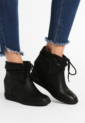 Pieds en jean bleu foncé à ourlet effiloché portant des bottines compensées noires avec fermetures éclair latérales et lacets à l’avant.