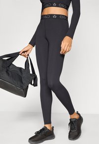 Leggings noirs avec une large ceinture arborant un logo, accompagnés d'un sac de sport noir et de chaussures de sport noires, présentant un design élégant et ajusté.