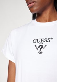 Bílý bavlněný tričko s černě vyšitým logem "GUESS U.S.A." s otazníkem a textem "WASHED JEANS" ve geometrickém designu.