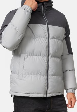 Homme portant une doudoune gris clair et gris foncé avec une fermeture éclair à l'avant et des poignets élastiques, tenant le col près de son menton.
