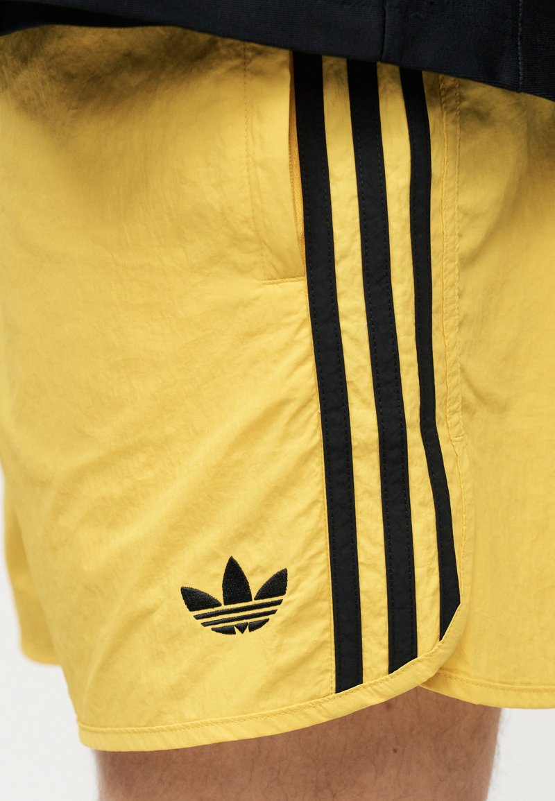Gule shorts med sorte Adidas tre striber og logo, båret af en person med synligt ben og sort topkant.