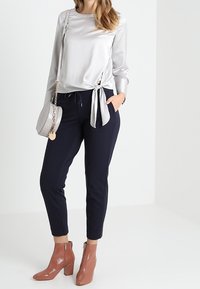 Blusa di raso argentato con dettaglio di nodo laterale, pantaloni tapered blu navy e stivaletti lucidi marroni con tacco a blocco. Piccola borsa rotonda argentata.