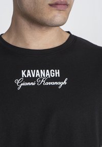 Musta puuvillapaita, jossa on pyöreä kaula-aukko, ja jossa on valkoista painettua tekstiä: "KAVANAGH" lihavoituna ja "Gianni Kavanagh" kursiivilla.
