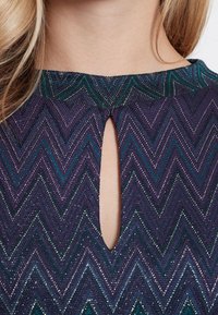 Top a motivo viola e teal con design a zig-zag, caratterizzato da un'apertura a forma di chiave nel collo e tessuto testurizzato con accenti metallici.