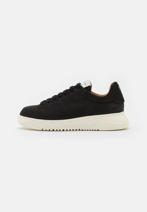 Must mustaga sneaker, mille valge kummist tald, ringjate paeltega, pehme kraega ning minimalismi stiilis. Sile tekstuur, madal profiil.