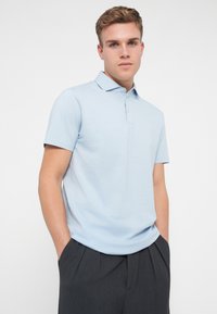 Gaiši zils polo krekls ar apkakli, īsām piedurknēm, standarta piegriezumu un smalku zīmola logo uz krūtīm; apvienots ar tumšām biksēm.