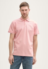 Helles rosa Poloshirt mit drei Knöpfen, einem Kragen und weißen Abschlüssen an den Ärmeln. Der Stoff wirkt weich mit einer feinen Textur.