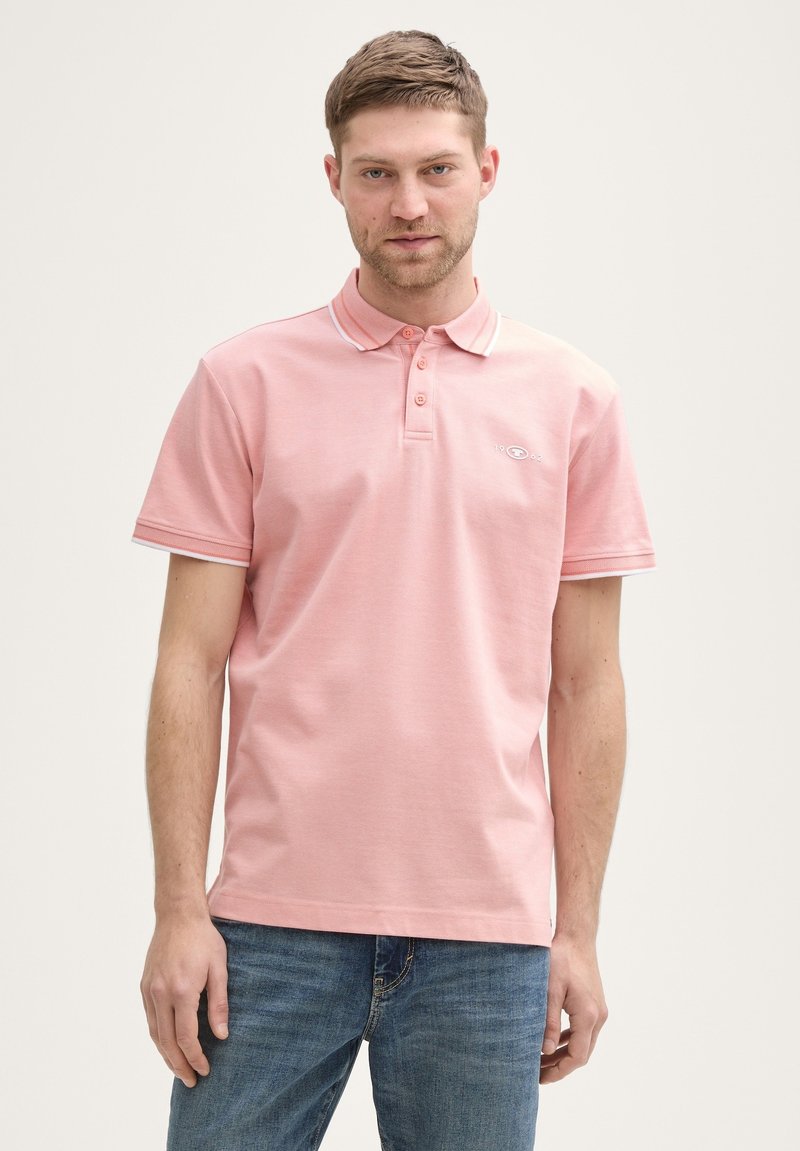 Helles rosa Poloshirt mit drei Knöpfen, einem Kragen und weißen Abschlüssen an den Ärmeln. Der Stoff wirkt weich mit einer feinen Textur.