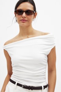 White off-shoulder top with ruched detailing, paired with black belt. Model wears brown-tinted sunglasses and silver earrings. Smooth texture.