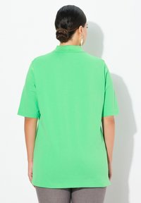 Zielona koszulka polo z krótkim rękawem, bawełniany materiał, luźny krój, jednolity kolor, płaski kołnierzyk, bez wzorów, noszona z dopasowanymi szarymi spodniami.