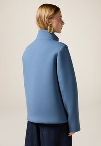 Giacca over-size blu realizzata in tessuto liscio, con colletto alto e spalle scese. Indossata con pantaloni in denim scuro.