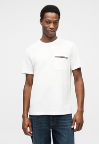 Λευκό βαμβακερό T-shirt με τσέπη στο στήθος που έχει κόκκινες και μπλε ρίγες, στρογγυλή λαιμόκοψη και κοντά μανίκια, συνδυασμένο με σκούρο μπλε τζιν.