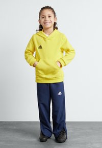 Felpa gialla con cappuccio e tasca frontale, con logo nero, abbinata a pantaloni tuta blu navy con fessure laterali e logo bianco, indossata con scarpe nere.