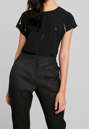 Femme portant un haut noir à manches courtes avec des rayures verticales en sequins et un pantalon noir texturé taille haute, sur fond gris uni.