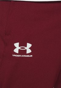 Burgundy träningsbyxor i strukturerad tyg. Har en vit Under Armour-logotyp på vänster sida nära midjan.