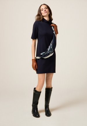 Vestido corto de manga corta en color navy con cuello simulado, combinado con guantes de cuero marrón y botas negras hasta la rodilla. Una bufanda estampada está colgada sobre el cuerpo.