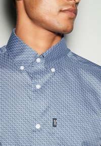 Camicia a motivo blu chiaro con colletto button-down, dotata di bottoni bianchi e di una piccola etichetta logo nera sul lato sinistro del petto.