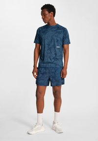 Man som står, klädd i matchande marinblå sporttröja och shorts, vita sneakers, tittar nedåt och åt vänster mot en enfärgad bakgrund.