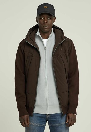 Bruine gewatteerde jas met een capuchon, voorzien van een grijze ritssluiting en een gestructureerde binnenlaag. Blauwe jeans en een zwarte pet maken de outfit compleet.
