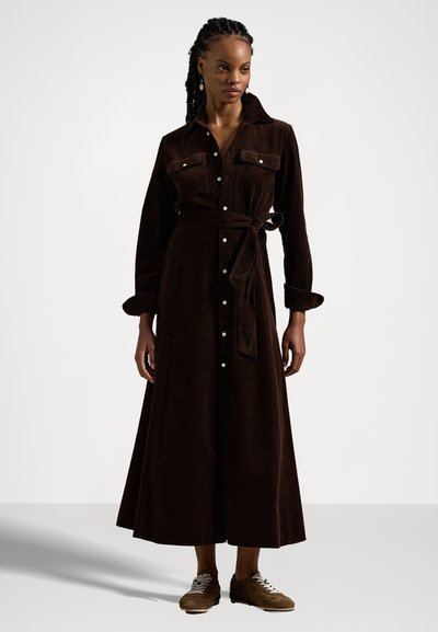 Brun kordfløjlskjole med knappet front, lange ærmer og bindebånd i taljen. Den har to brystlommer og en flydende, ankel-lang nederdel.