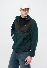 Fleecepaita, jossa tummanvihreitä ja mustia paneeleja, oransseja yksityiskohtia, vetoketjulliset taskut ja korkea kaulus. Yhdistetty sinisiin farkkuihin ja valkoiseen lippikseen.