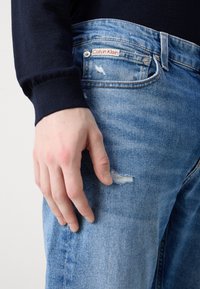 Svijetloplave traperice s detaljima strgane na bedru, ukrašene crvenom oznakom Calvin Klein na pojasu.