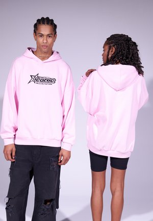 Hellrosa Oversized-Hoodie mit schwarzem Logo, ausgestattet mit einer Kordelzugkapuze und gerippten Bündchen, kombiniert mit stark verwaschenen schwarzen Jeans und Shorts.