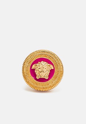 Bouton rond en or avec des bords striés, présentant un emblème de tête de Méduse dorée sur un fond circulaire rose vif.