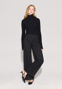 Czarny sweter z golfem o prążkowanej teksturze, połączony z wysokim stanem czarnych szerokich spodni i wypolerowanymi czarnymi szpilkami, na szarym tle.